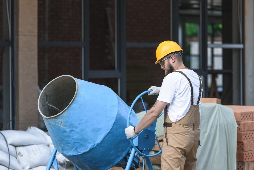 construction worker in hardhat and protective googles working with concrete mixer on construction